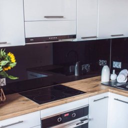 Duo Serwis - Nowoczesna kuchnia z białymi szafkami, drewnianym blatem i czarnym panelem szklanym. Widoczna płyta indukcyjna, piekarnik i dekoracje. Ujęcie z góry.