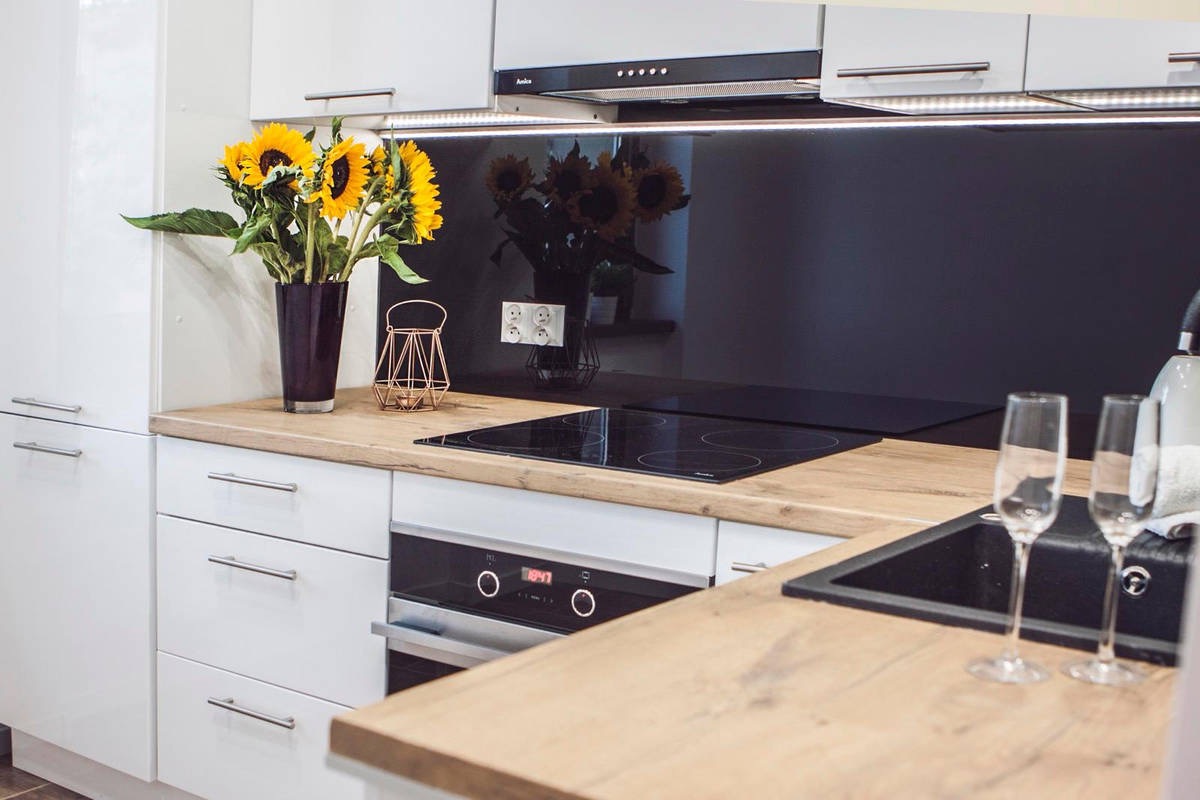 Nowoczesna kuchnia z białymi szafkami, drewnianym blatem i czarnym panelem szklanym. Na blacie wazon ze słonecznikami i dekoracje. Ujęcie z perspektywy.