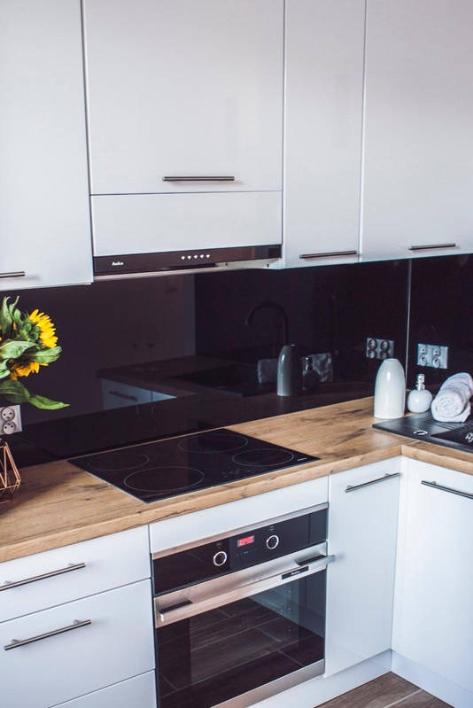 Nowoczesna kuchnia z białymi szafkami, drewnianym blatem i czarnym panelem szklanym. Widoczna płyta indukcyjna, piekarnik i dekoracje. Ujęcie z góry.