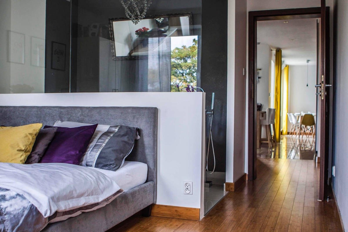 Nowoczesna sypialnia z łazienką oddzieloną szklaną ścianą, widok na parkiet i jadalnię przez otwarte drzwi. Minimalistyczny design, stonowana kolorystyka z akcentami fioletu i żółci.