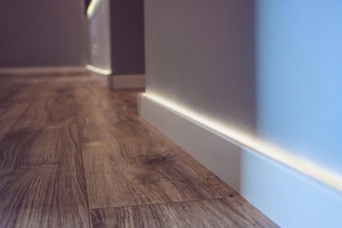 Drewnopodobne panele podłogowe i białe listwy przypodłogowe z subtelnym podświetleniem LED wzdłuż ściany. Nowoczesne wykończenie wnętrza z bliska.