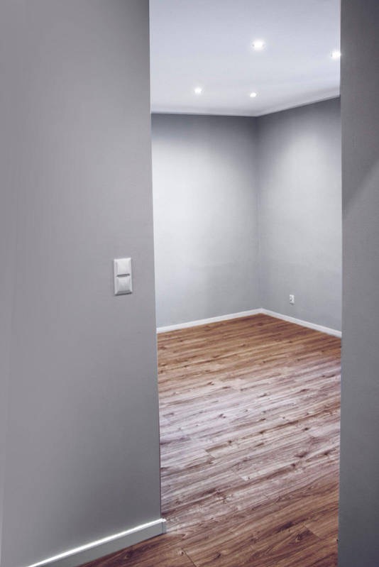 Szare ściany i drewnopodobna podłoga w pustym pokoju z oświetleniem sufitowym. Widok z korytarza, z włącznikiem światła na ścianie po lewej.