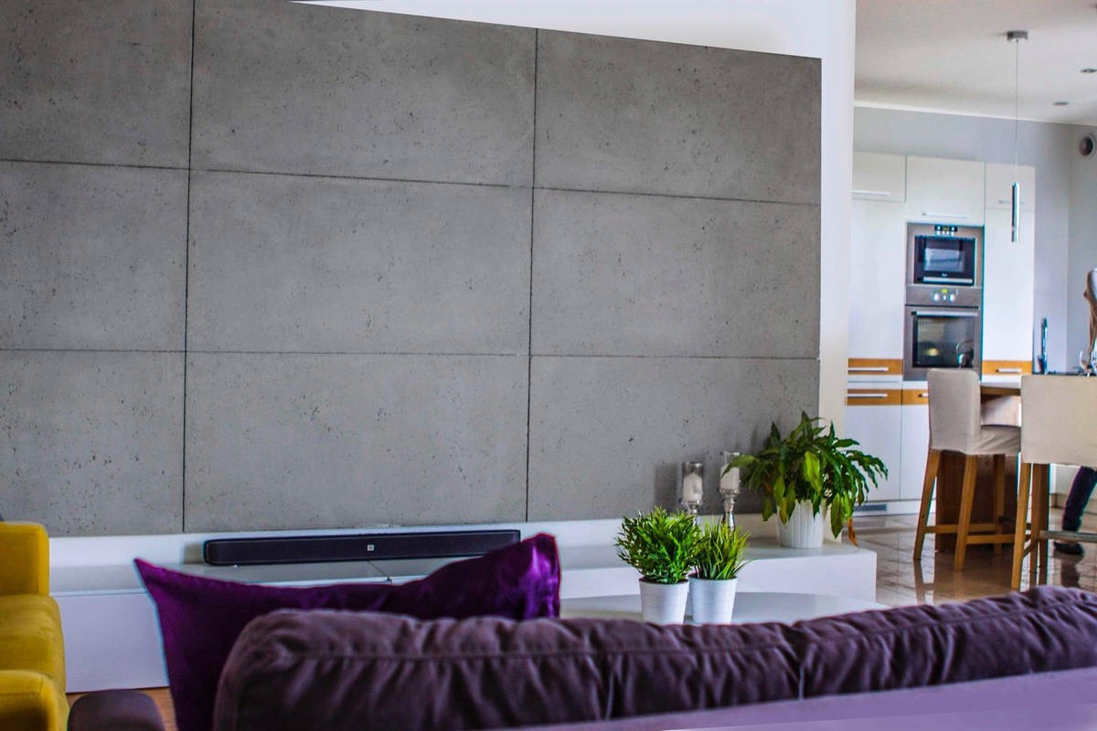 Nowoczesny salon z szarą ścianą wykończoną betonowymi panelami, minimalistycznym oświetleniem i elementami dekoracyjnymi. Widok na otwartą kuchnię w tle.