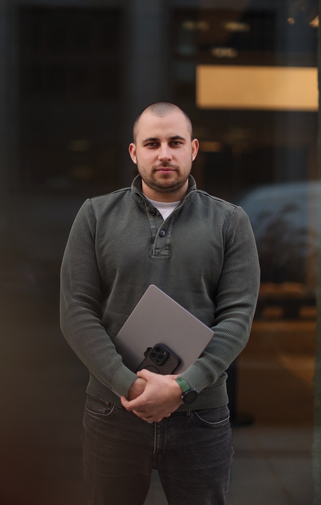 Młody mężczyzna w zielonej bluzie trzyma laptop i telefon na tle szklanej fasady budynku. Widoczny zegarek na ręce i ciemne jeansy.