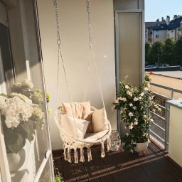 Kuba Fiej - Przytulny balkon z huśtawką hamakową, poduszkami i kwiatami w donicach. Ciemne deski podłogowe i widok na budynki w tle. Słoneczny dzień.