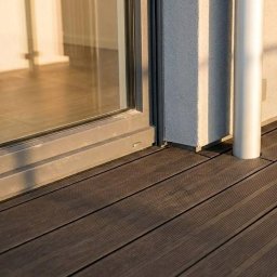 Kuba Fiej - Ciemnobrązowe deski tarasowe przy szarej ramie okiennej i białej rurze spustowej w słoneczny dzień. Widok z niskiego kąta.