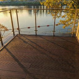 Kuba Fiej - Nowoczesny taras z brązowych desek z widokiem na rzekę i zabudową ze szkła. Jesienne drzewa w tle. Ujęcie z poziomu podłogi.