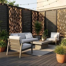 Kuba Fiej - Nowoczesny taras z deskami w odcieniach szarości, otoczony dekoracyjnym, ażurowym panelem i roślinami w donicach. Meble wypoczynkowe i nastrojowe oświetlenie.