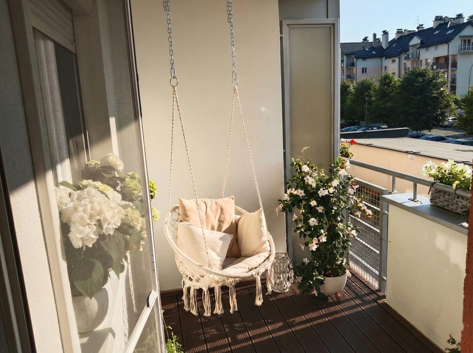 Przytulny balkon z huśtawką hamakową, poduszkami i kwiatami w donicach. Ciemne deski podłogowe i widok na budynki w tle. Słoneczny dzień.