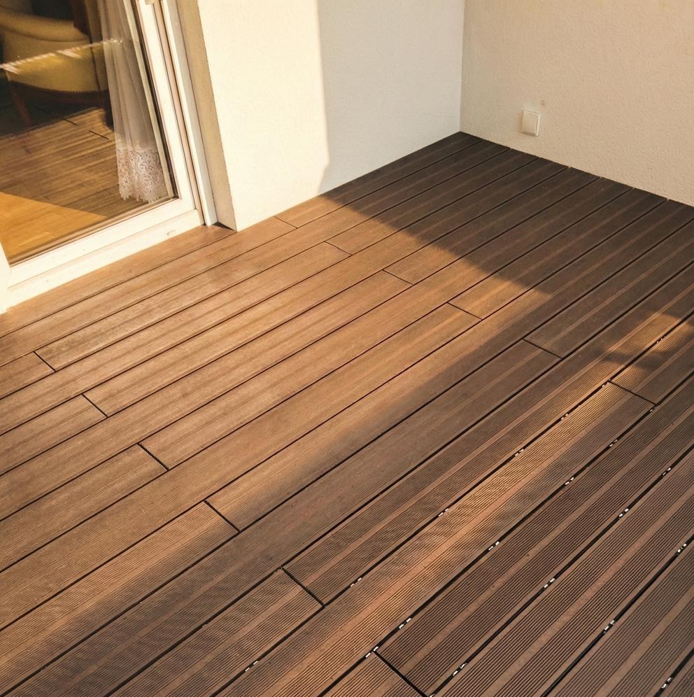 Balkon z brązową podłogą z desek kompozytowych, oświetlony ciepłym światłem słonecznym wpadającym przez okno balkonowe. Widoczna faktura desek i biała ściana.