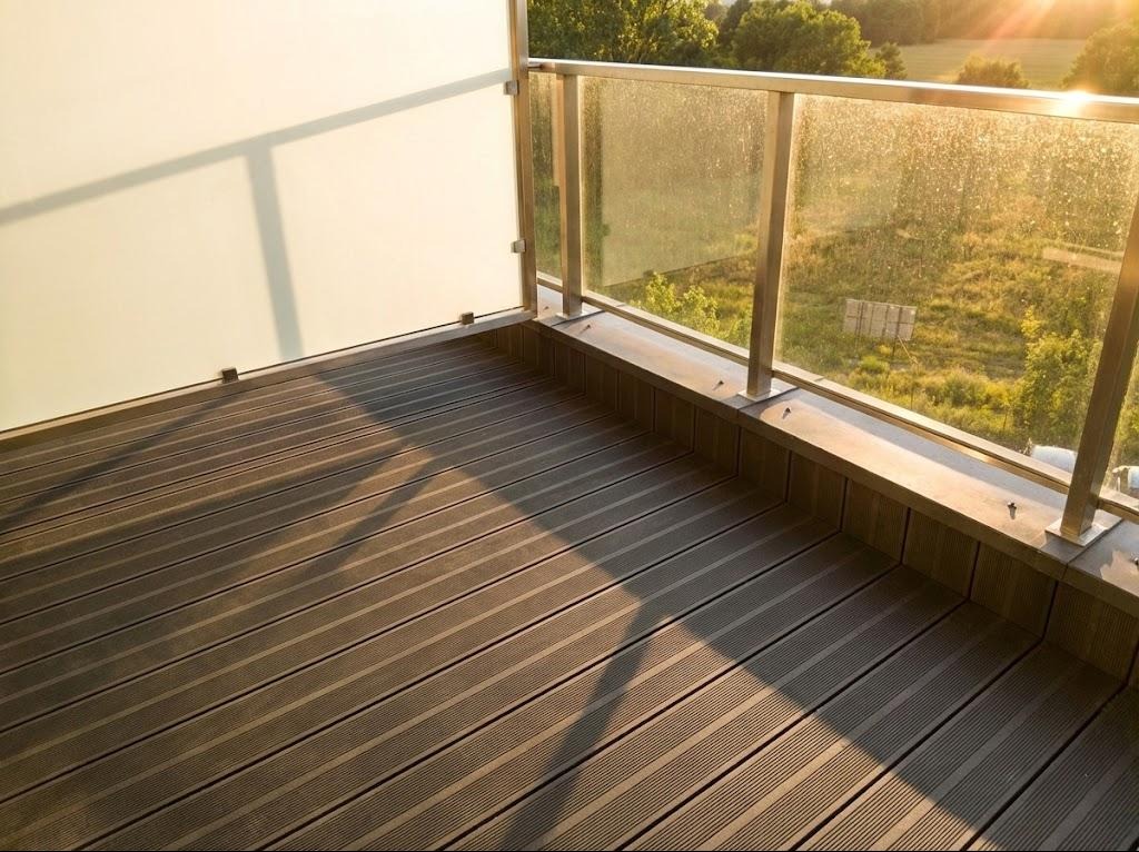 Nowoczesny balkon z brązową podłogą z desek kompozytowych, szklaną balustradą i widokiem na zieleń w tle, oświetlony ciepłym, popołudniowym słońcem.