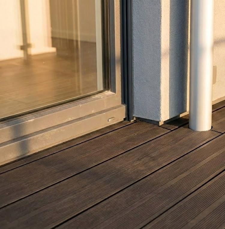 Ciemnobrązowe deski tarasowe przy szarej ramie okiennej i białej rurze spustowej w słoneczny dzień. Widok z niskiego kąta.