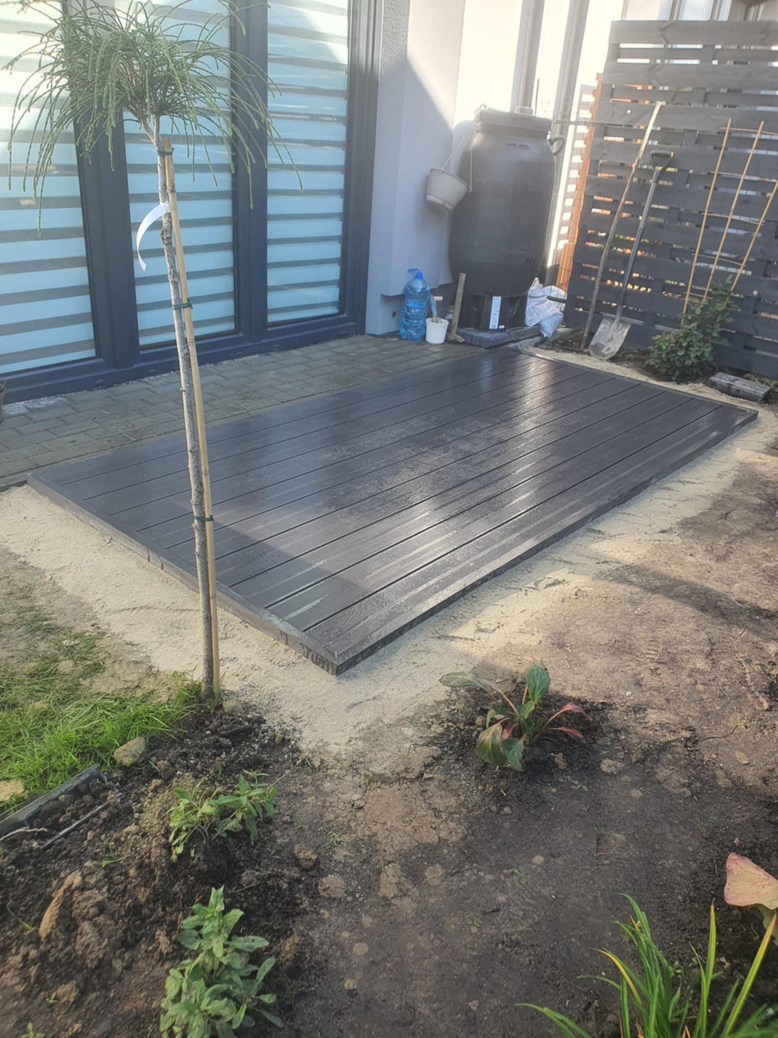 Nowy, ciemnoszary taras kompozytowy w trakcie budowy, otoczony piaskiem i ziemią. W tle widoczna ściana budynku z oknami i drewniane ogrodzenie.