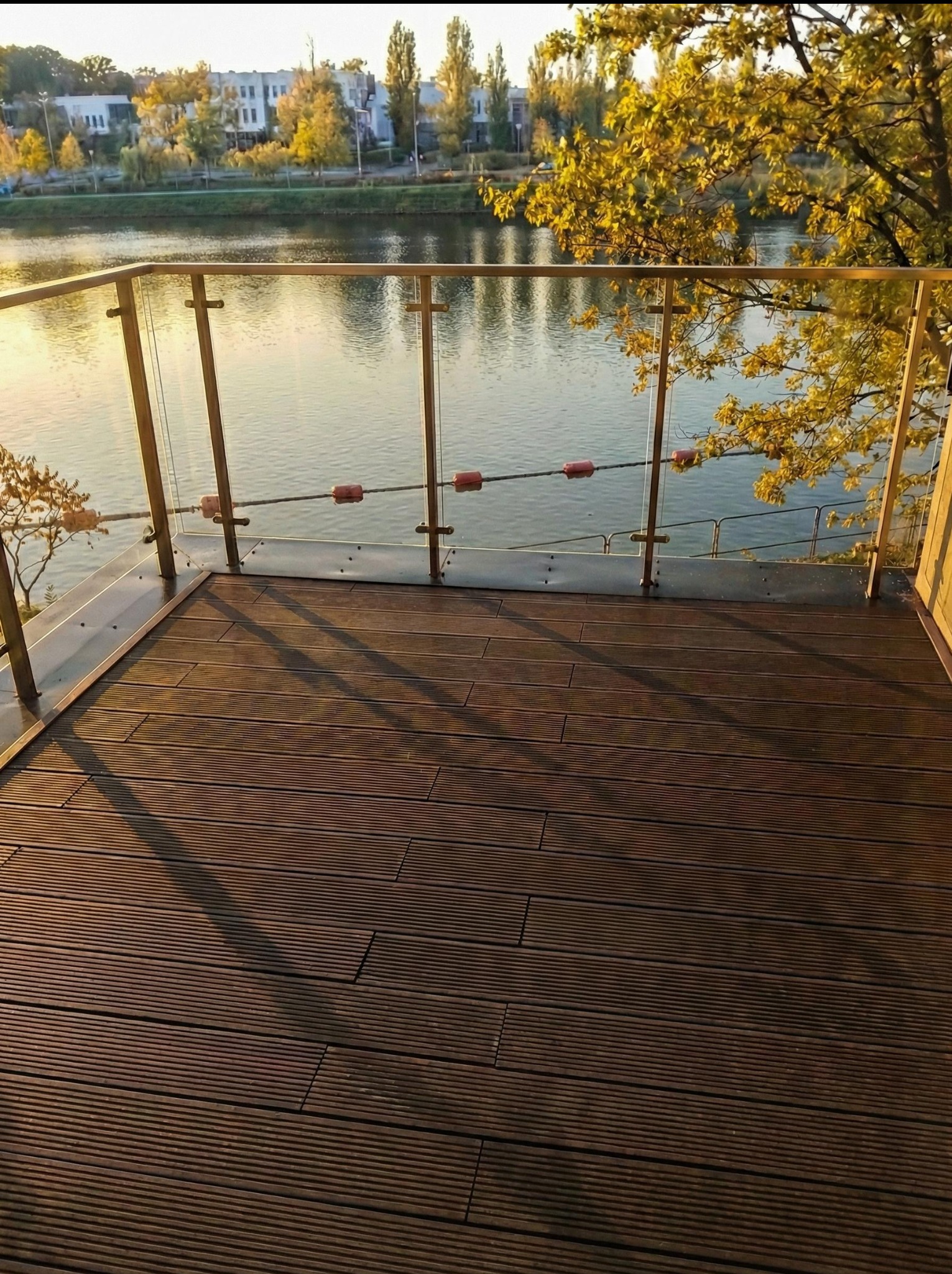 Nowoczesny taras z brązowych desek z widokiem na rzekę i zabudową ze szkła. Jesienne drzewa w tle. Ujęcie z poziomu podłogi.