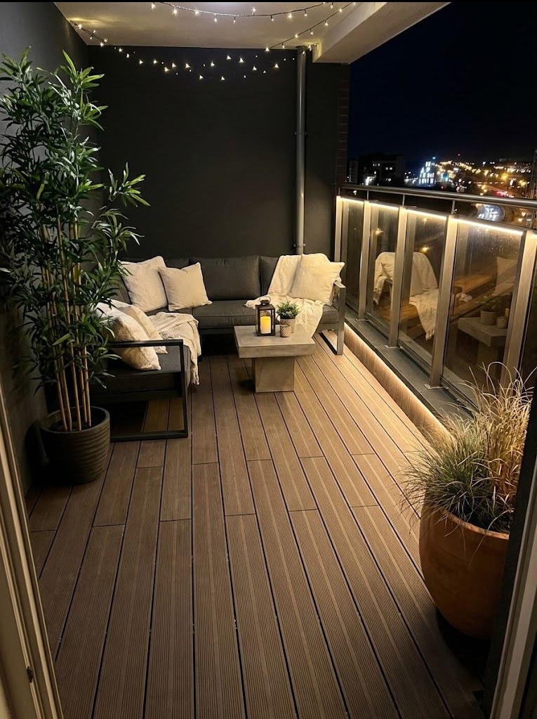 Przytulny balkon nocą: deski w odcieniach brązu, szara sofa z poduszkami, lampion na stoliku i dekoracyjne oświetlenie LED wzdłuż balustrady.