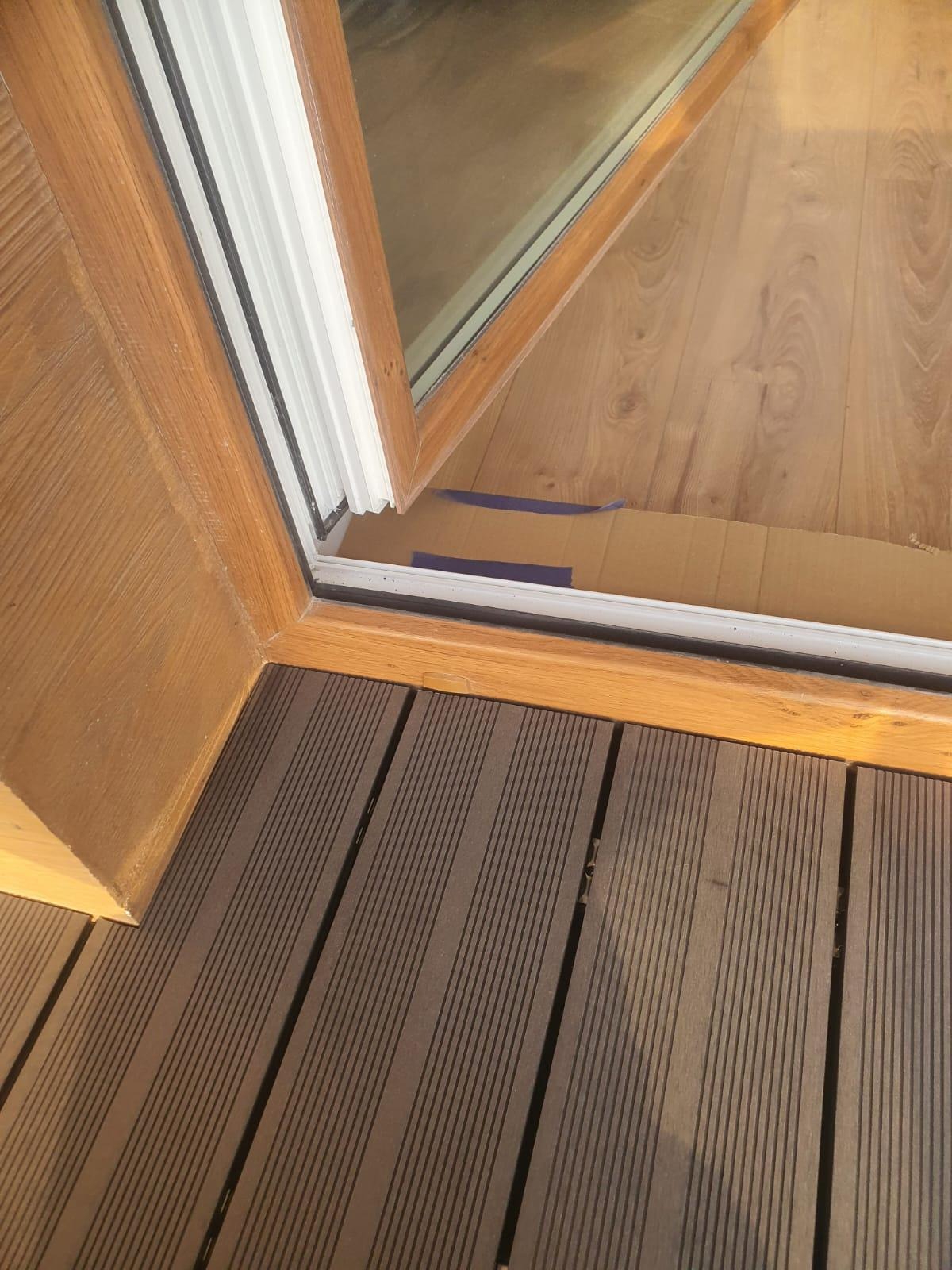 Fragment balkonu z brązową deską kompozytową i drewnianą ramą okienną. Widoczne zabezpieczenia pod oknem. Ujęcie z góry, bliski kadr.