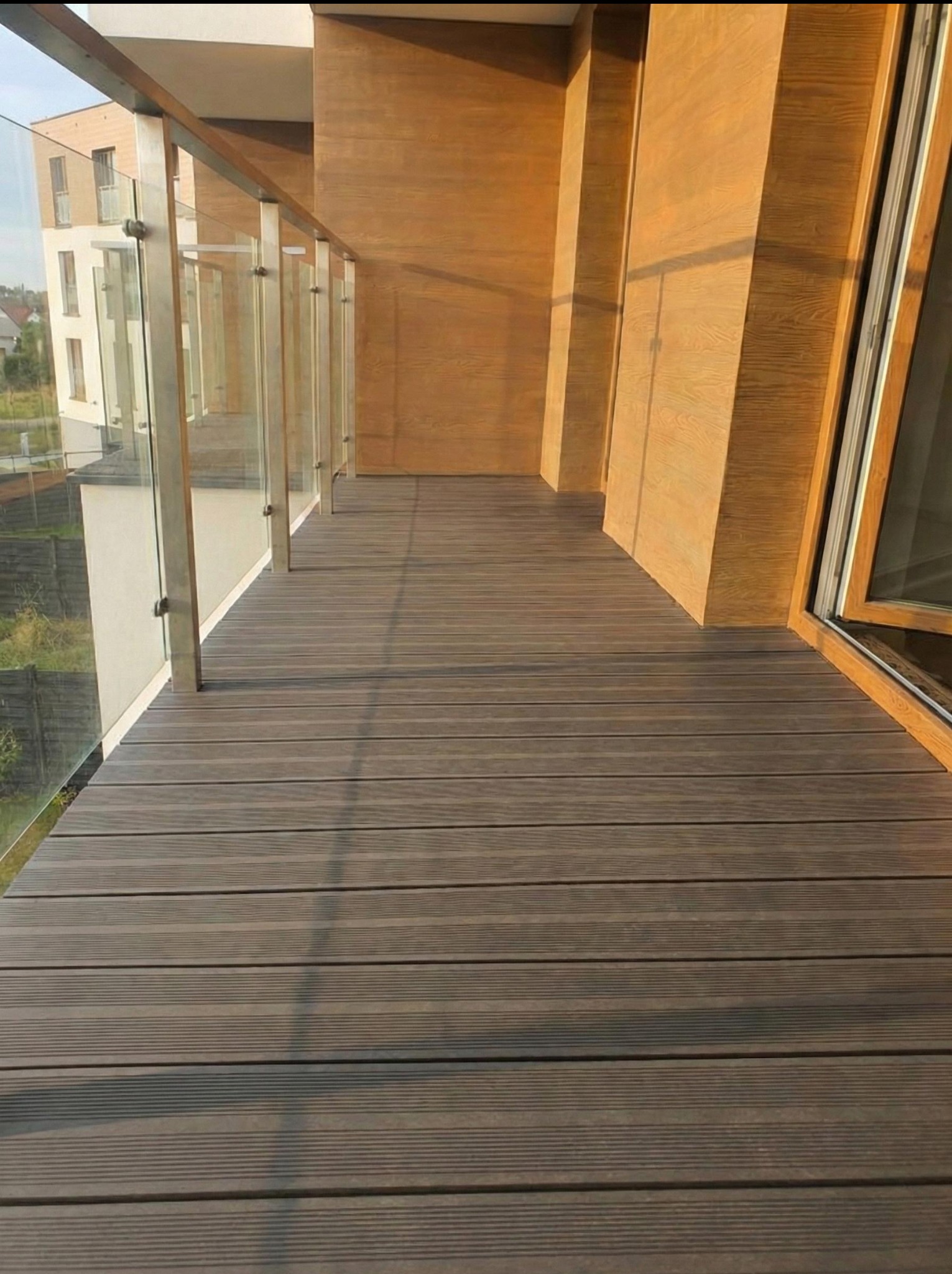 Nowoczesny balkon z szarą podłogą z desek kompozytowych i szklaną balustradą. Ściana wykończona panelami imitującymi drewno. Widok na budynek mieszkalny.
