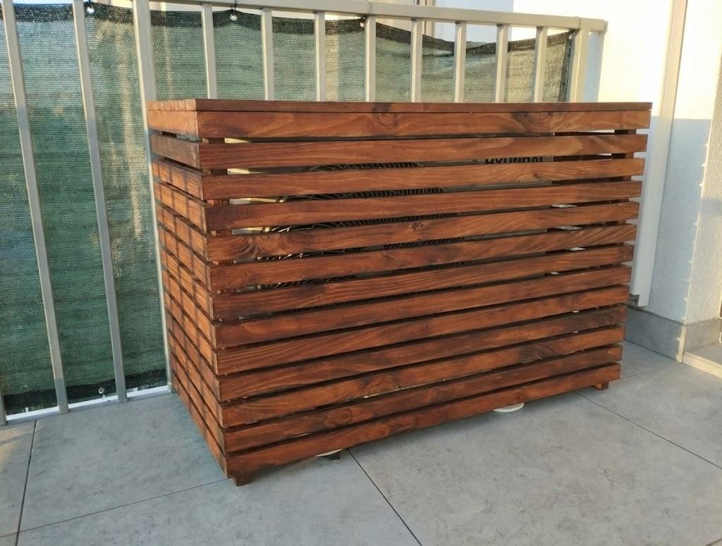 Drewniana osłona na klimatyzator na balkonie, wykonana z bejcowanego drewna w ciepłym odcieniu brązu. Widoczne poziome deski z przerwami, tworzące ażurową konstrukcję.