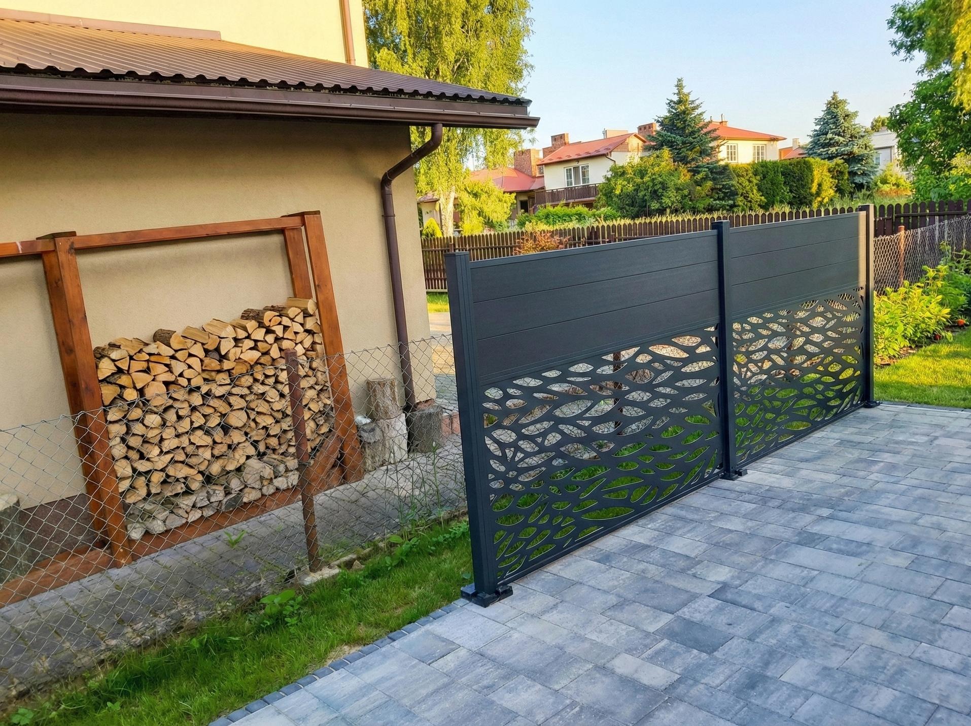 Nowoczesny, ażurowy panel ogrodzeniowy w kolorze grafitowym, oddzielający posesję. W tle widoczny skład drewna i domy sąsiadów.