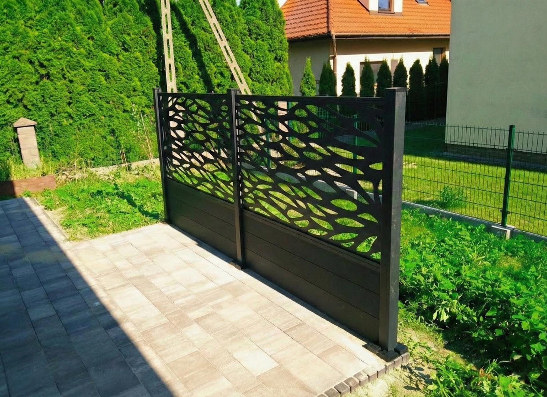 Nowoczesna, ażurowa przesłona z ciemnego kompozytu, oddzielająca taras z jasnej kostki od zielonego ogrodu. W tle dom z czerwonym dachem i wysokie tuje.
