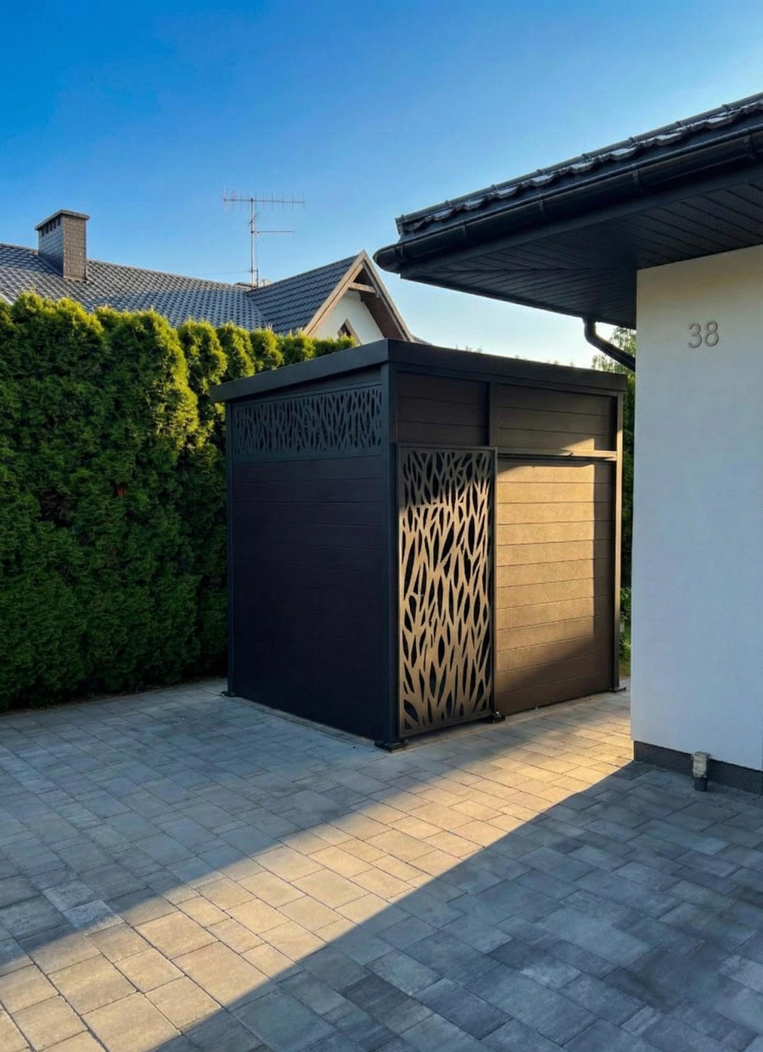 Nowoczesna, czarna zabudowa z ażurowym panelem, przylegająca do białej ściany budynku. Widoczny fragment kostki brukowej i zielony żywopłot w tle.