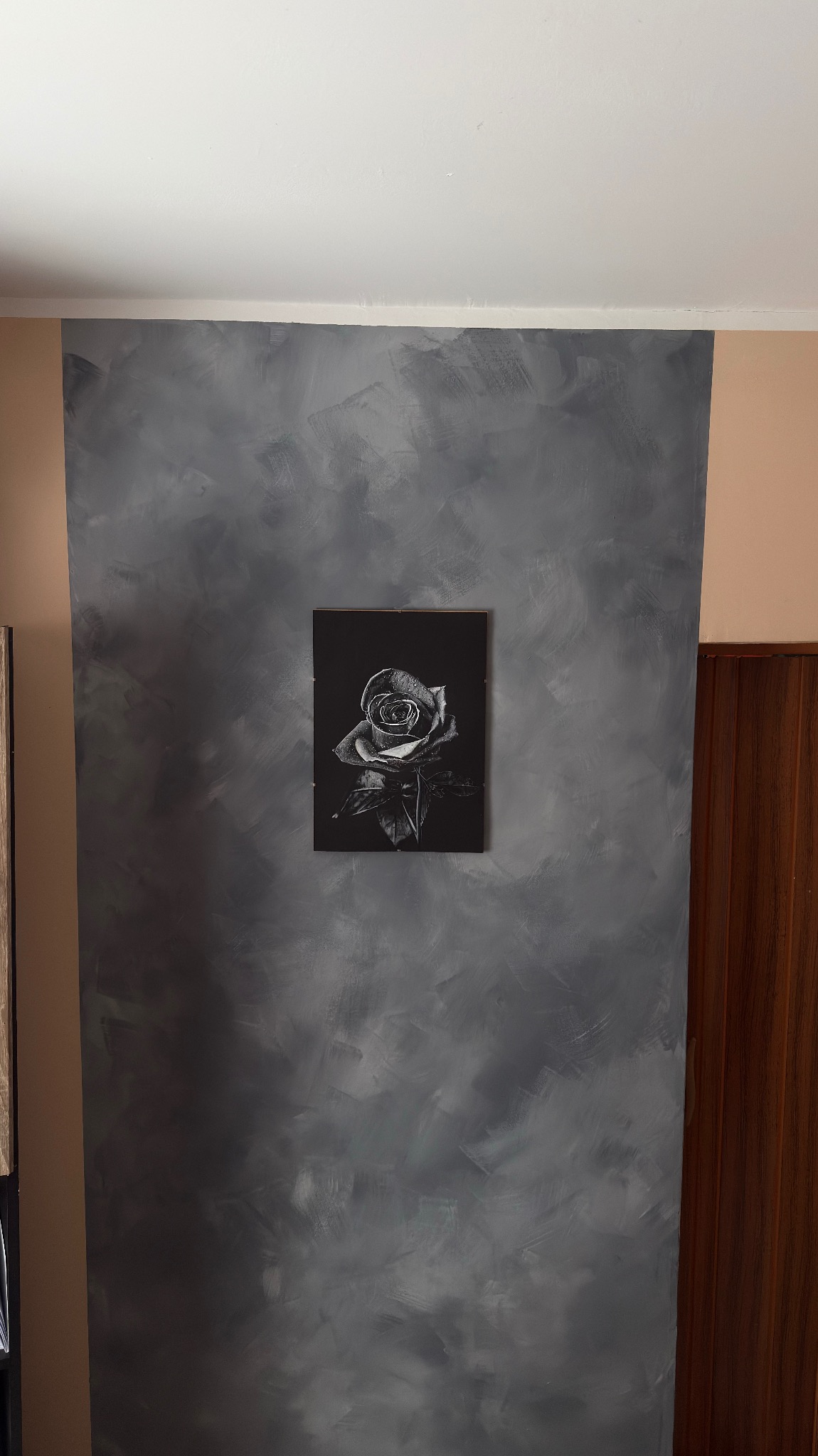 Ściana z dekoracyjną szarą fakturą, na której wisi czarno-biały obraz z motywem róży. Widoczny fragment brązowych drzwi po prawej stronie.