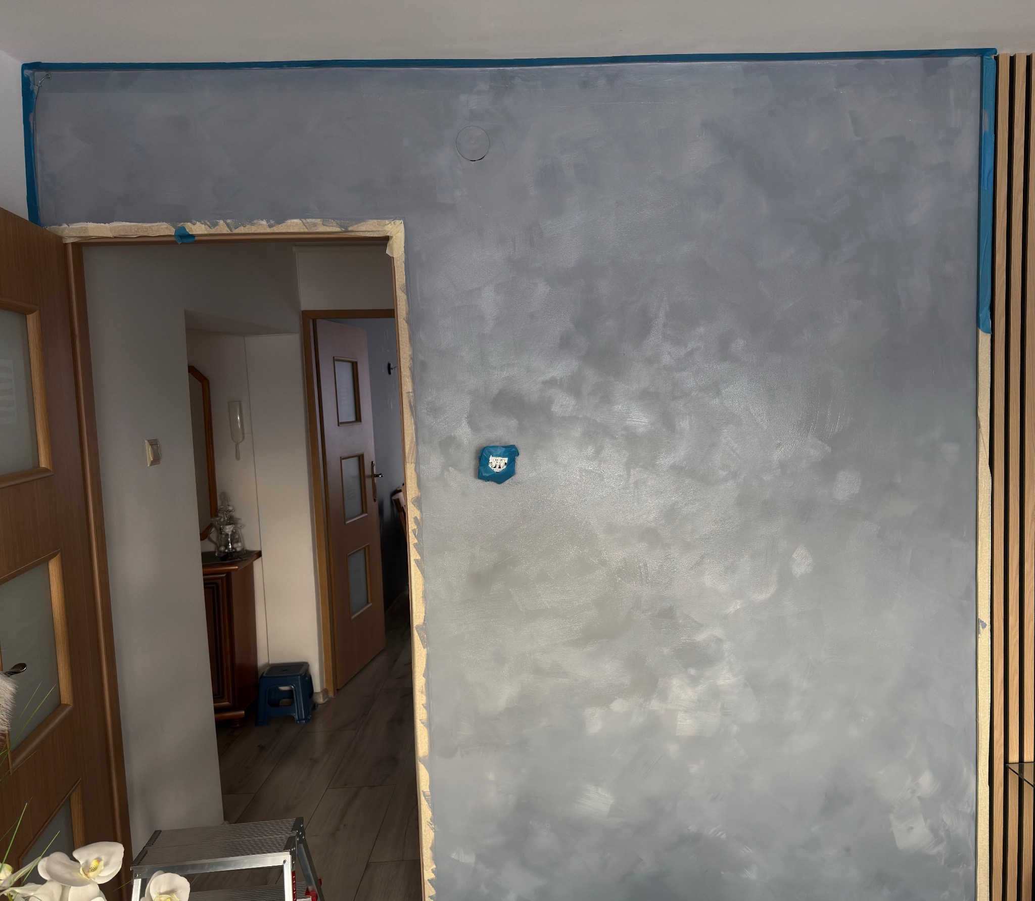 Ściana po dekoracyjnym malowaniu w odcieniach szarości w Bochni. Widoczne listwy drewniane i zabezpieczone taśmą malarską krawędzie.