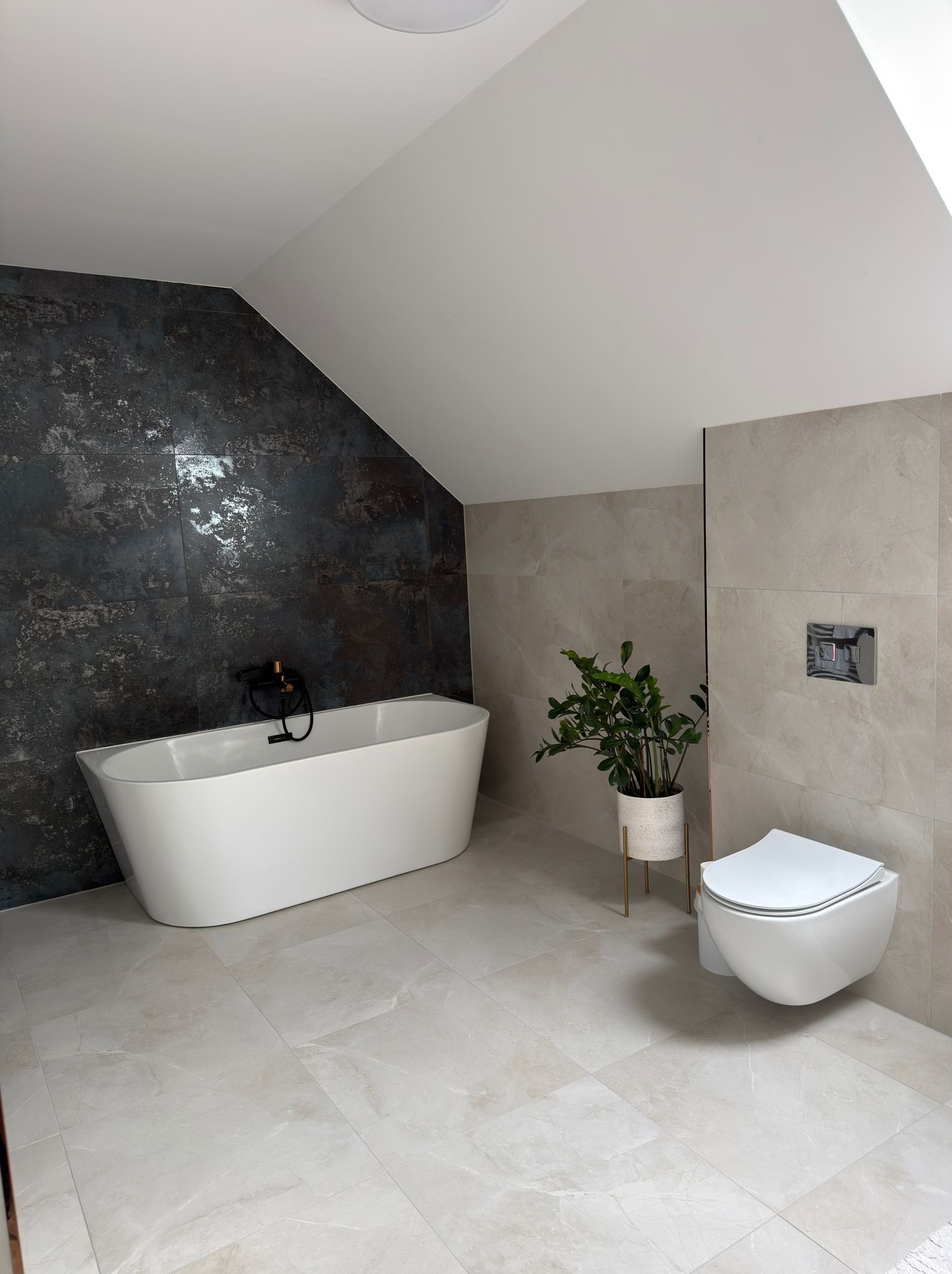 Nowoczesna łazienka na poddaszu z wolnostojącą wanną, ciemną ścianą z efektem rdzy i beżowymi płytkami. Uwagę przyciąga wisząca toaleta i roślina w ozdobnej doniczce.