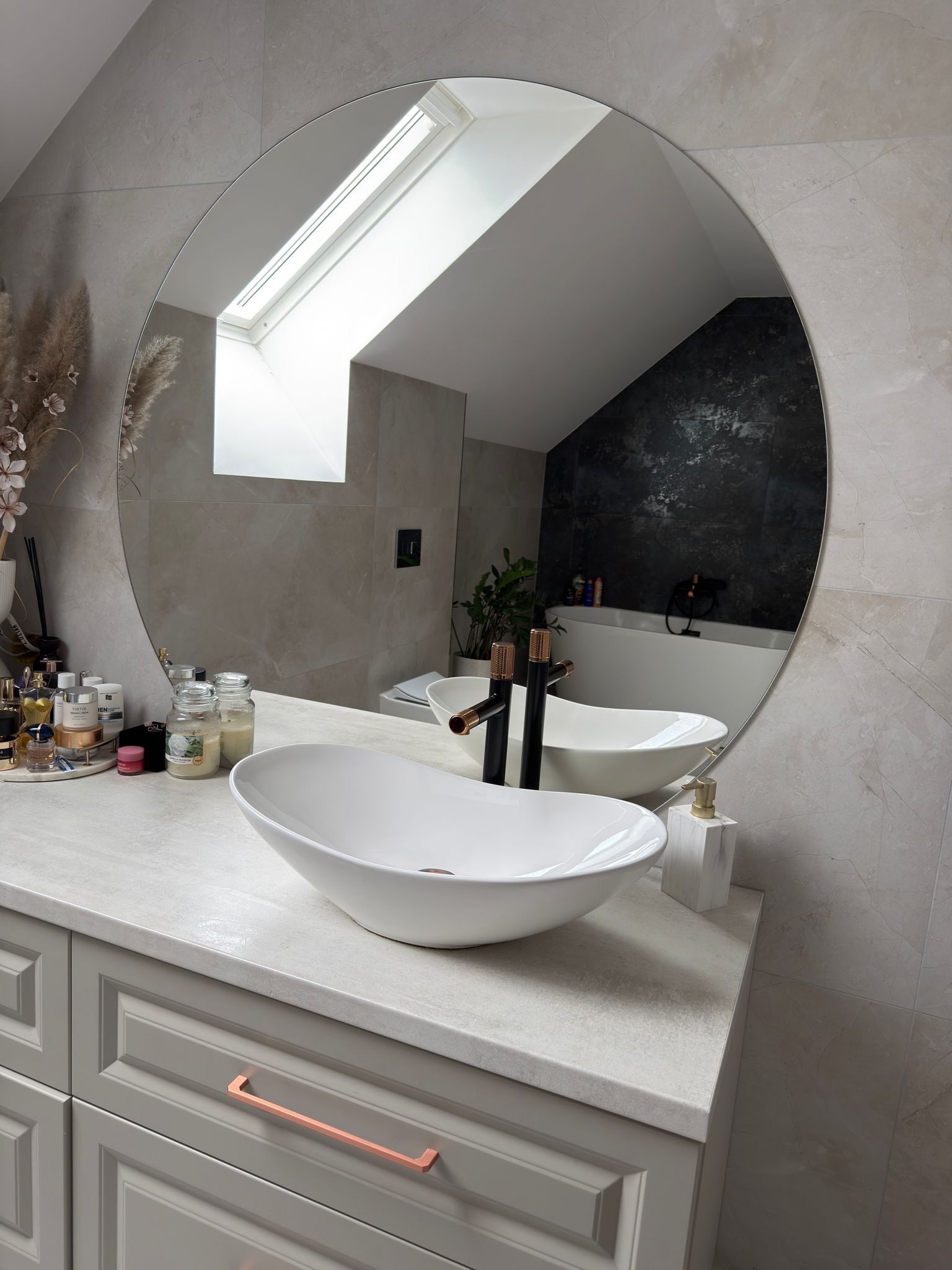 Stylowa łazienka z owalną umywalką nablatową, armaturą w kolorze miedzi i okrągłym lustrem, w którym odbija się okno dachowe i wanna. Jasne płytki imitujące marmur.