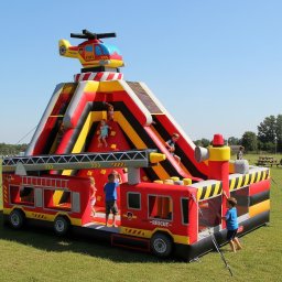 Nikolandia - Kolorowy dmuchany plac zabaw w kształcie wozu strażackiego z helikopterem na szczycie, dzieci bawią się na zjeżdżalni i ściance wspinaczkowej na tle zielonej trawy.