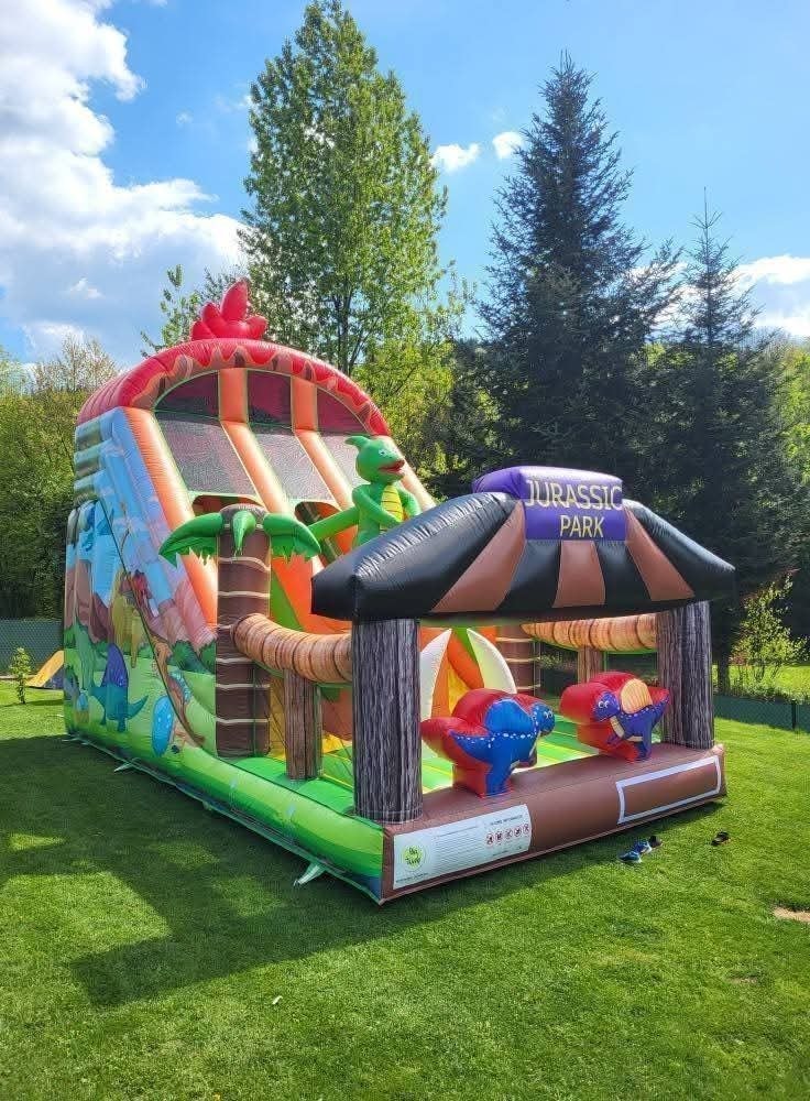 Kolorowy dmuchany zamek Jurassic Park z motywem dinozaurów, zjeżdżalnią i figurką dinozaura na tle zielonego trawnika i drzew.
