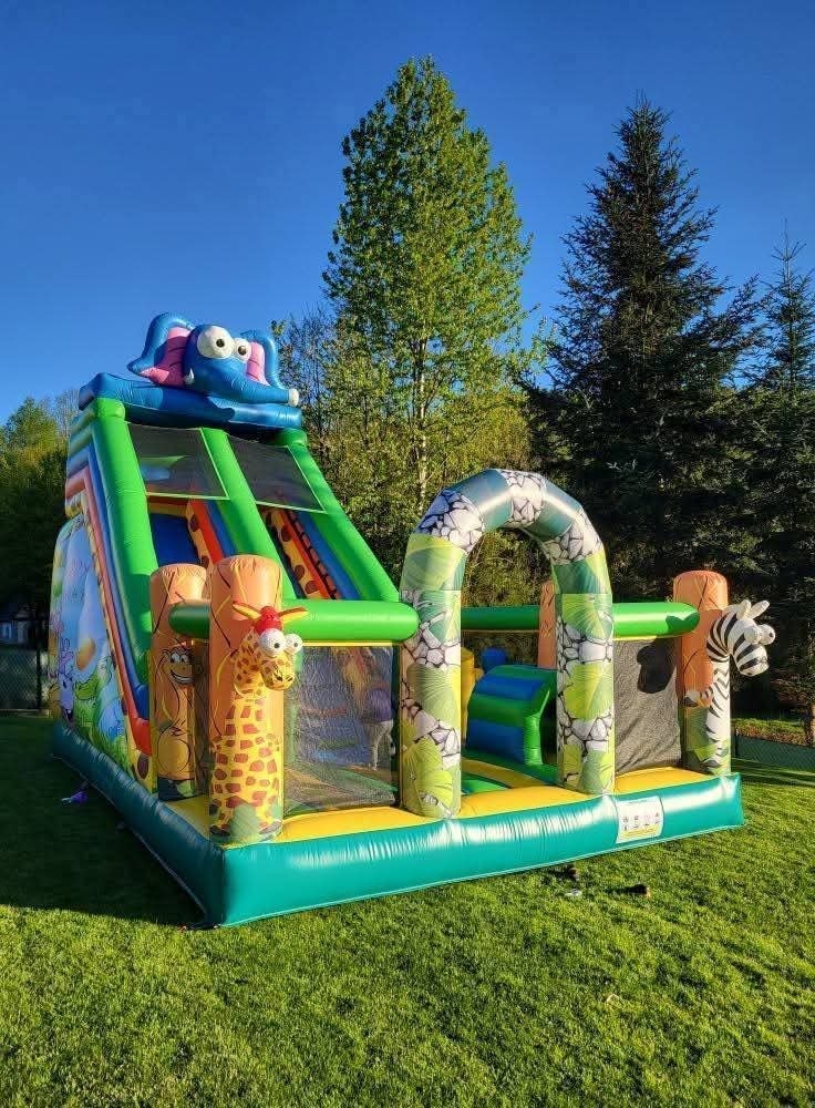 Kolorowy dmuchany zamek ze zjeżdżalnią i motywami zwierząt, ustawiony na trawniku w słoneczny dzień. Widoczne drzewa w tle.