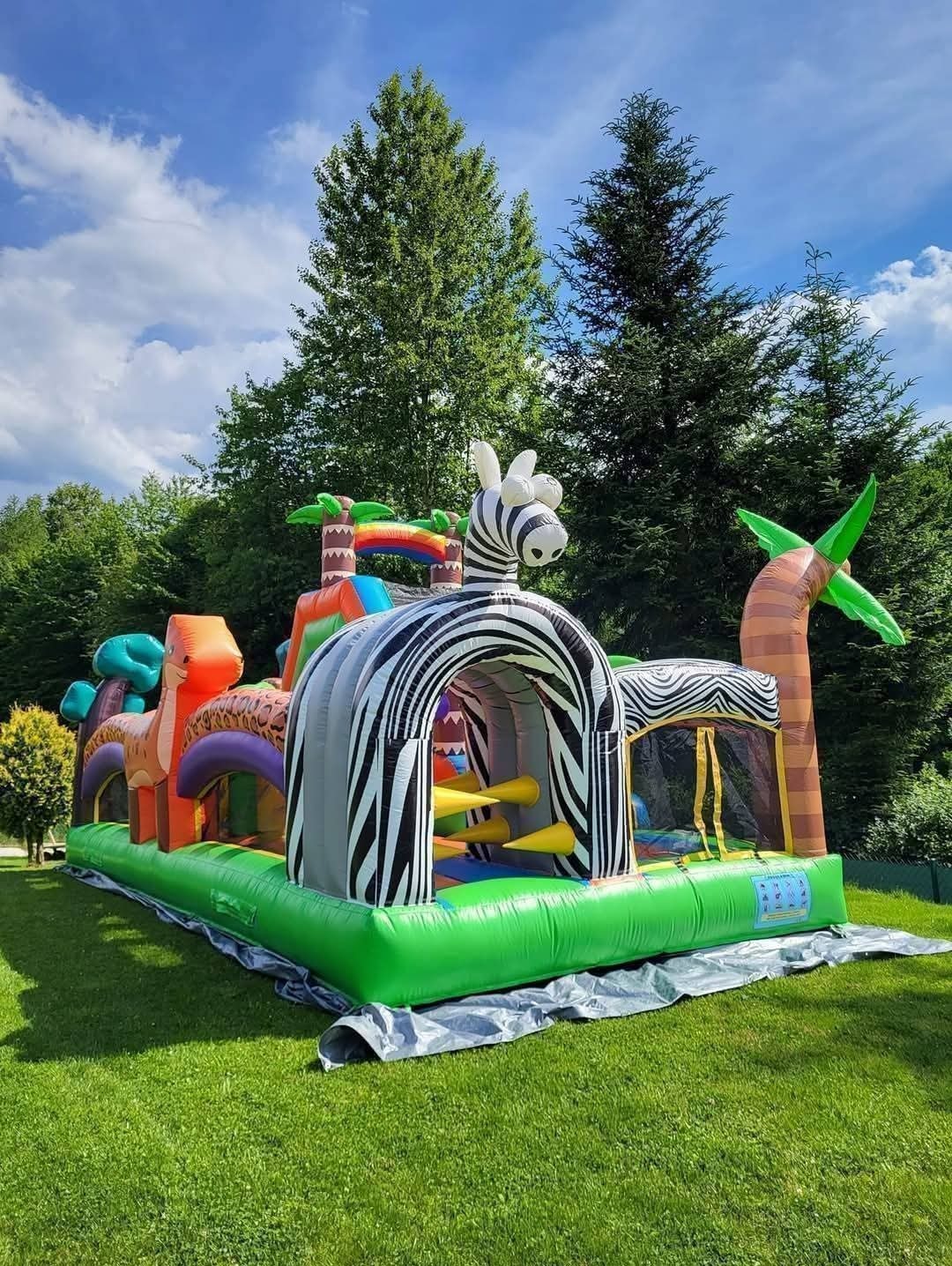 Kolorowy dmuchany plac zabaw z motywem zebry i dinozaurów na trawie, idealny na imprezy plenerowe dla dzieci. Widoczne drzewa w tle i błękitne niebo.