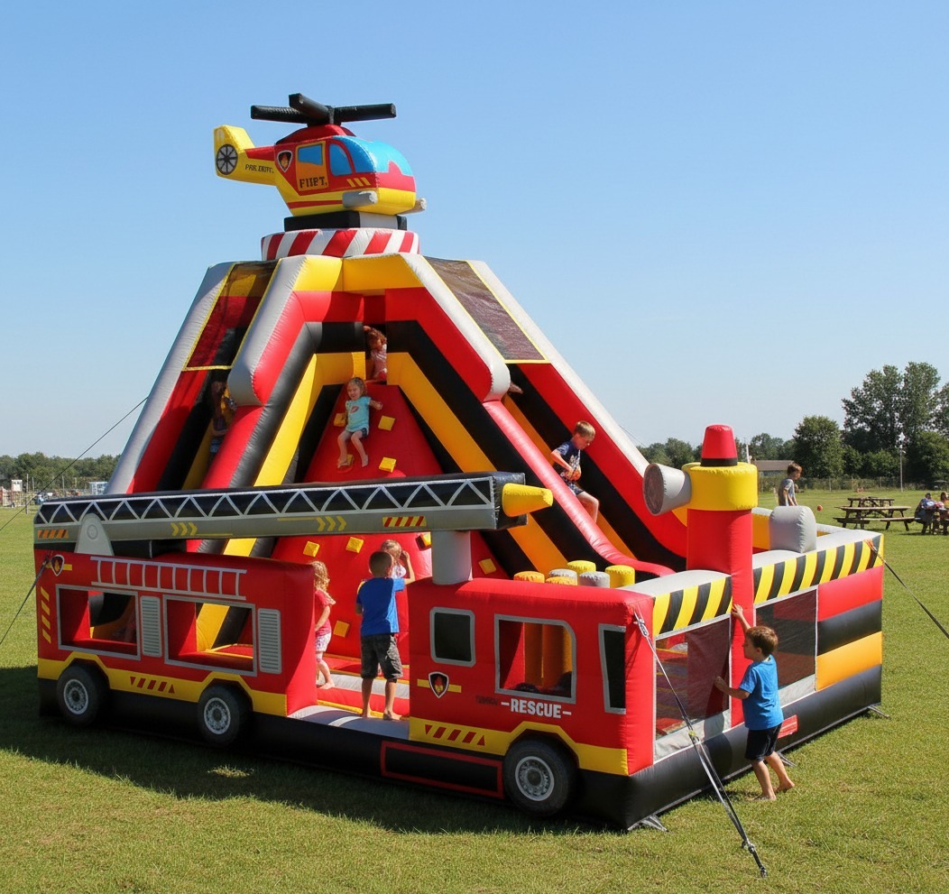 Kolorowy dmuchany plac zabaw w kształcie wozu strażackiego z helikopterem na szczycie, dzieci bawią się na zjeżdżalni i ściance wspinaczkowej na tle zielonej trawy.