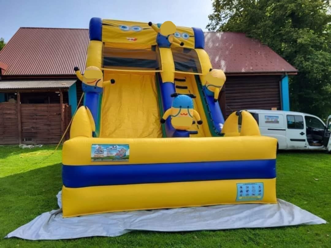 Żółto-niebieski dmuchany zamek ze zjeżdżalnią, stylizowany na Minionki, ustawiony na trawie przed budynkiem z czerwonym dachem i białym vanem w tle.