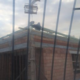 Kraw-Render - Budowa domu z cegły widziana przez kratę. Robotnicy na dachu montują konstrukcję, wspomagani przez dźwig. W tle pochmurne niebo.