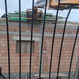 Kraw-Render - Widok z kabiny ładowarki teleskopowej na budynek w trakcie budowy. Żółta ładowarka podnosi drewniane elementy konstrukcyjne na mur z czerwonej cegły.