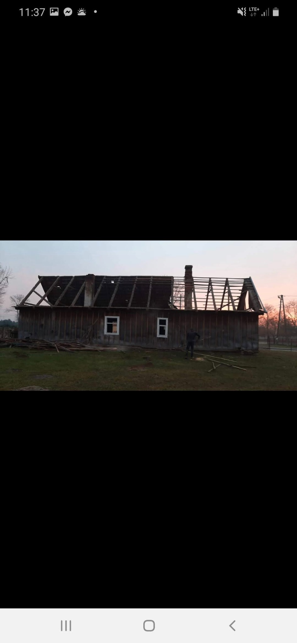 Demontaż dachu starego, drewnianego domu z kominem. Widoczne krokwie i pracownik na tle zachodzącego słońca. Częściowa rozbiórka budynku.