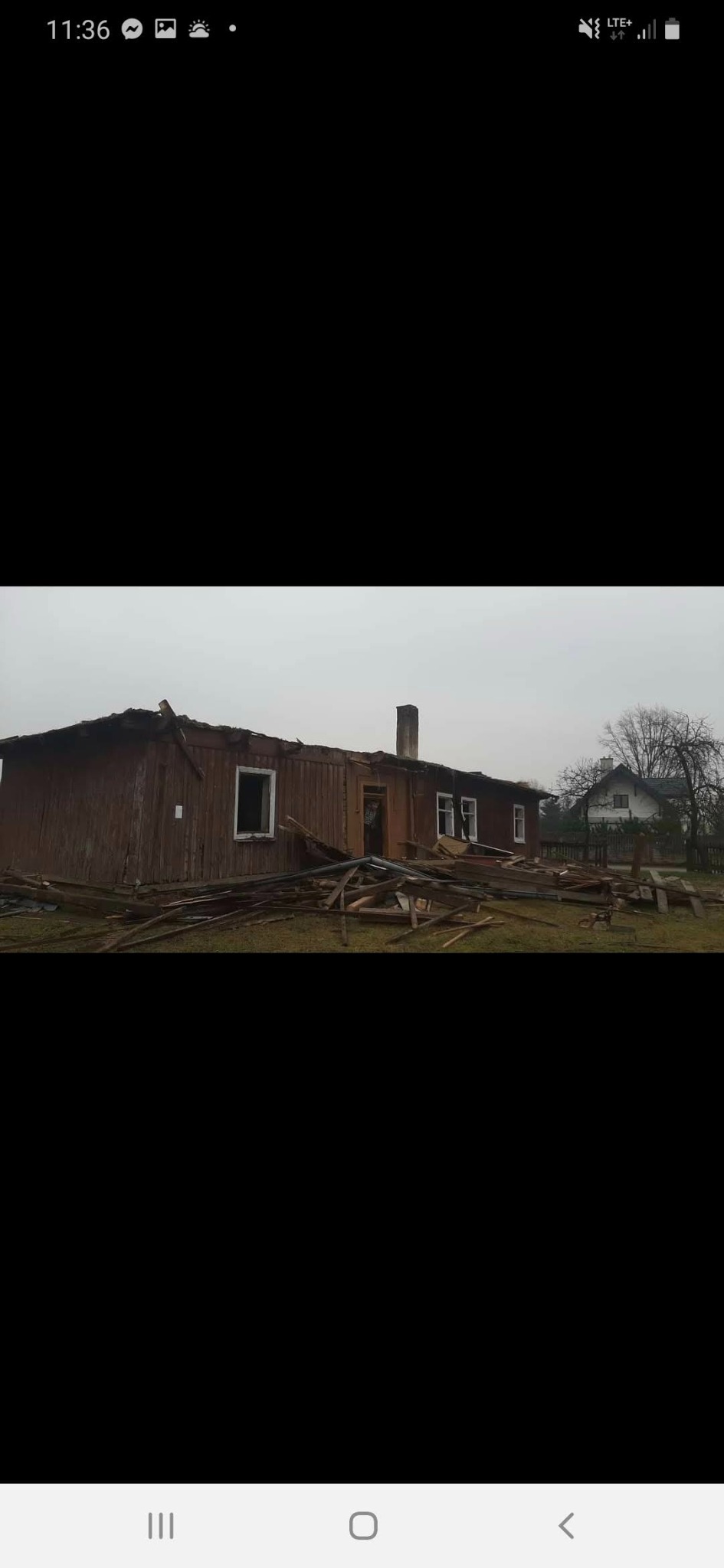 Uszkodzony, częściowo rozebrany, parterowy budynek z drewnianą elewacją. Wokół porozrzucane elementy konstrukcyjne. W tle widoczny inny budynek i drzewa.