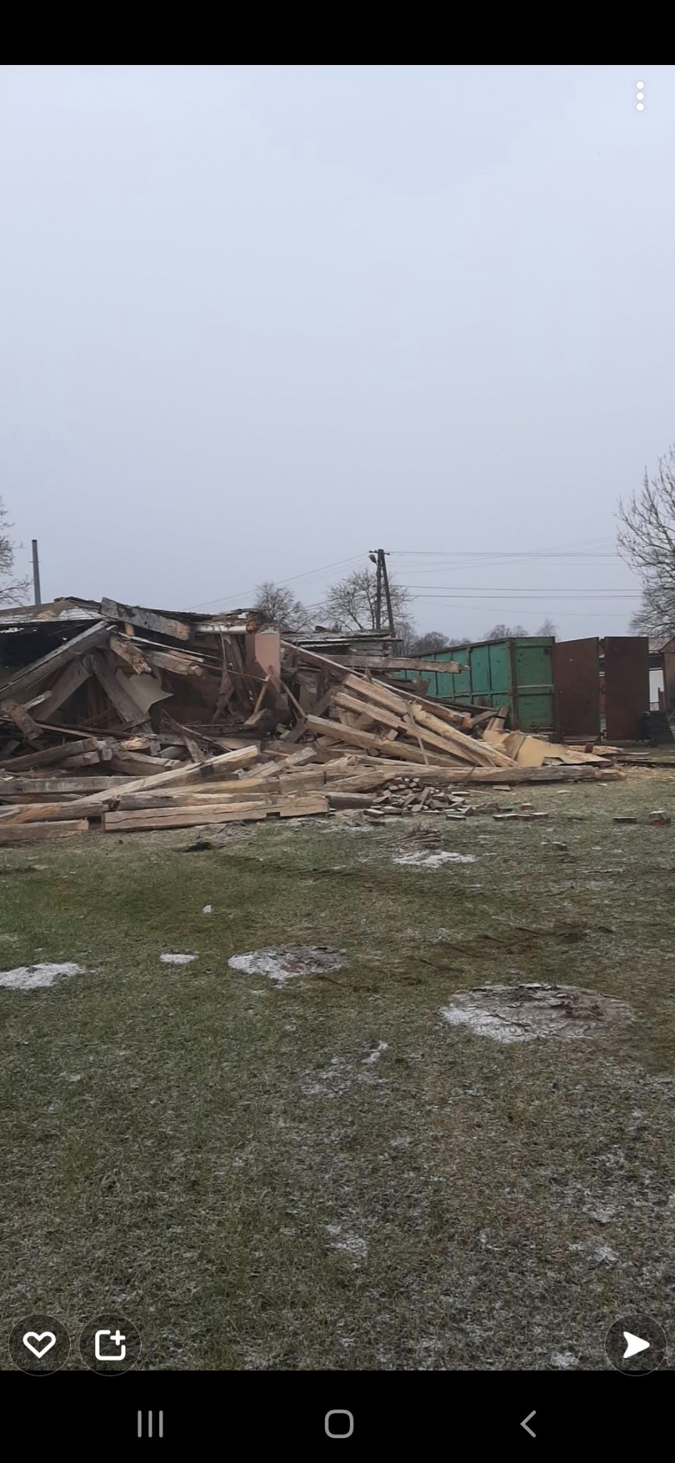 Sterta gruzu i drewnianych belek po rozbiórce budynku, z zielonym kontenerem w tle, na zewnątrz w pochmurny dzień. Widoczne ślady śniegu na trawie.