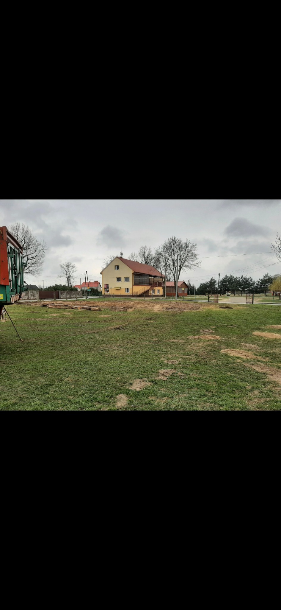 Przygotowany teren pod budowę domu jednorodzinnego. Widoczne ślady niwelacji, w tle budynek mieszkalny i fragment koparki. Pochmurny dzień.