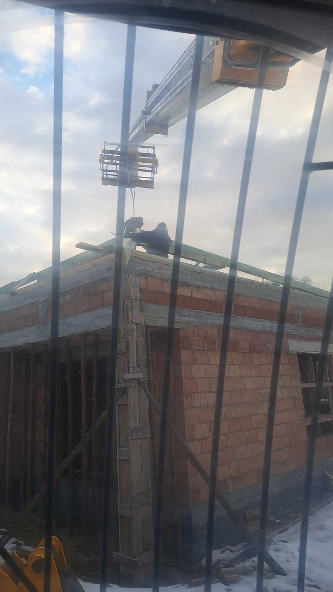 Budowa domu z cegły widziana przez kratę. Robotnicy na dachu montują konstrukcję, wspomagani przez dźwig. W tle pochmurne niebo.