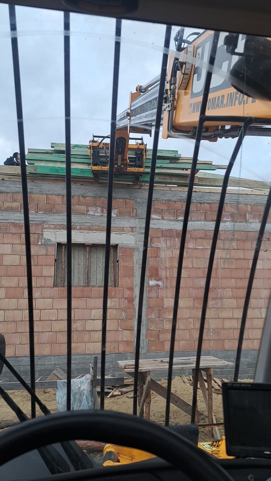 Widok z kabiny ładowarki teleskopowej na budynek w trakcie budowy. Żółta ładowarka podnosi drewniane elementy konstrukcyjne na mur z czerwonej cegły.