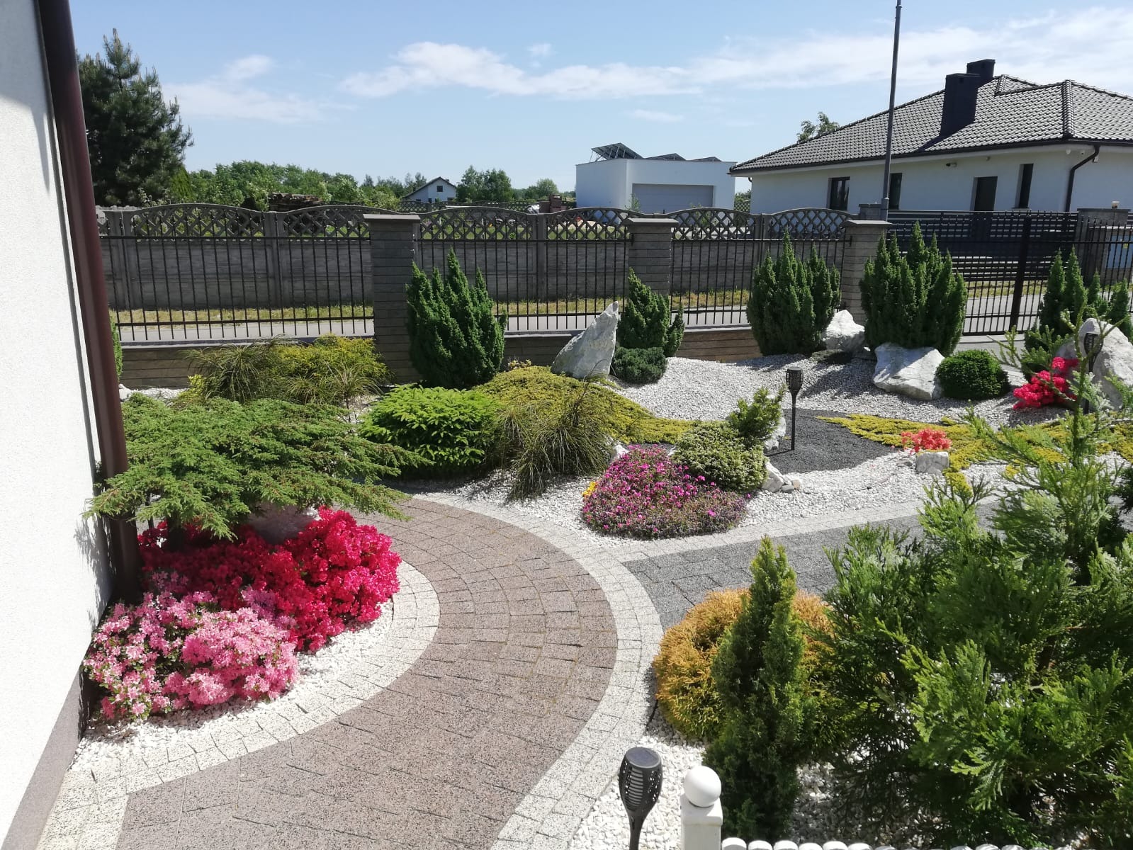 Ogród z krzewami, kamieniami i ścieżką z kostki brukowej. Widoczne różowe azalie, tuje i ozdobne kamienie. W tle dom z ciemną dachówką i ogrodzenie.