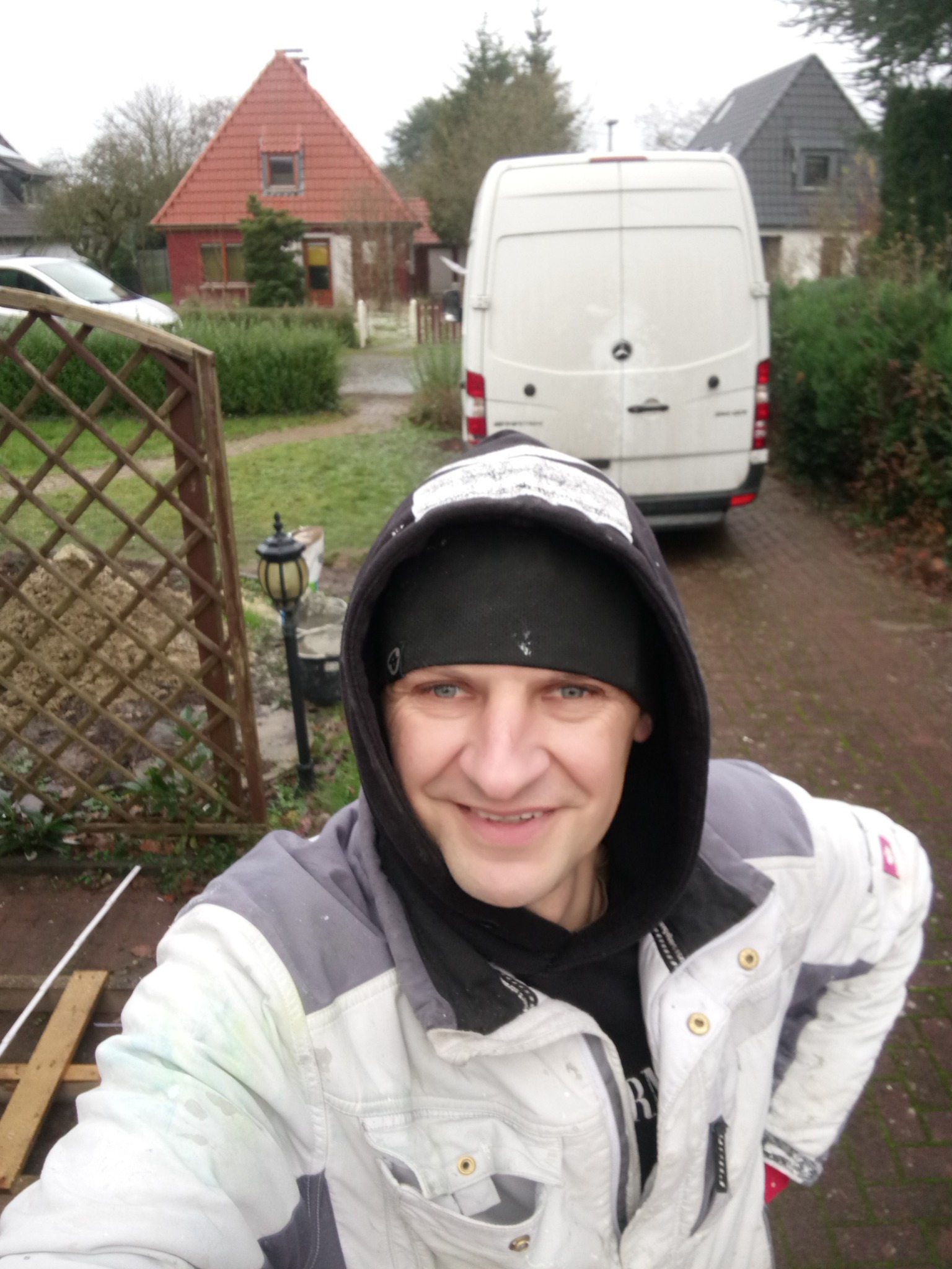 Uśmiechnięty mężczyzna w roboczej kurtce z plamami farby, na tle białego busa i domu z czerwoną dachówką. Widoczny fragment drewnianego płotu i lampa ogrodowa.