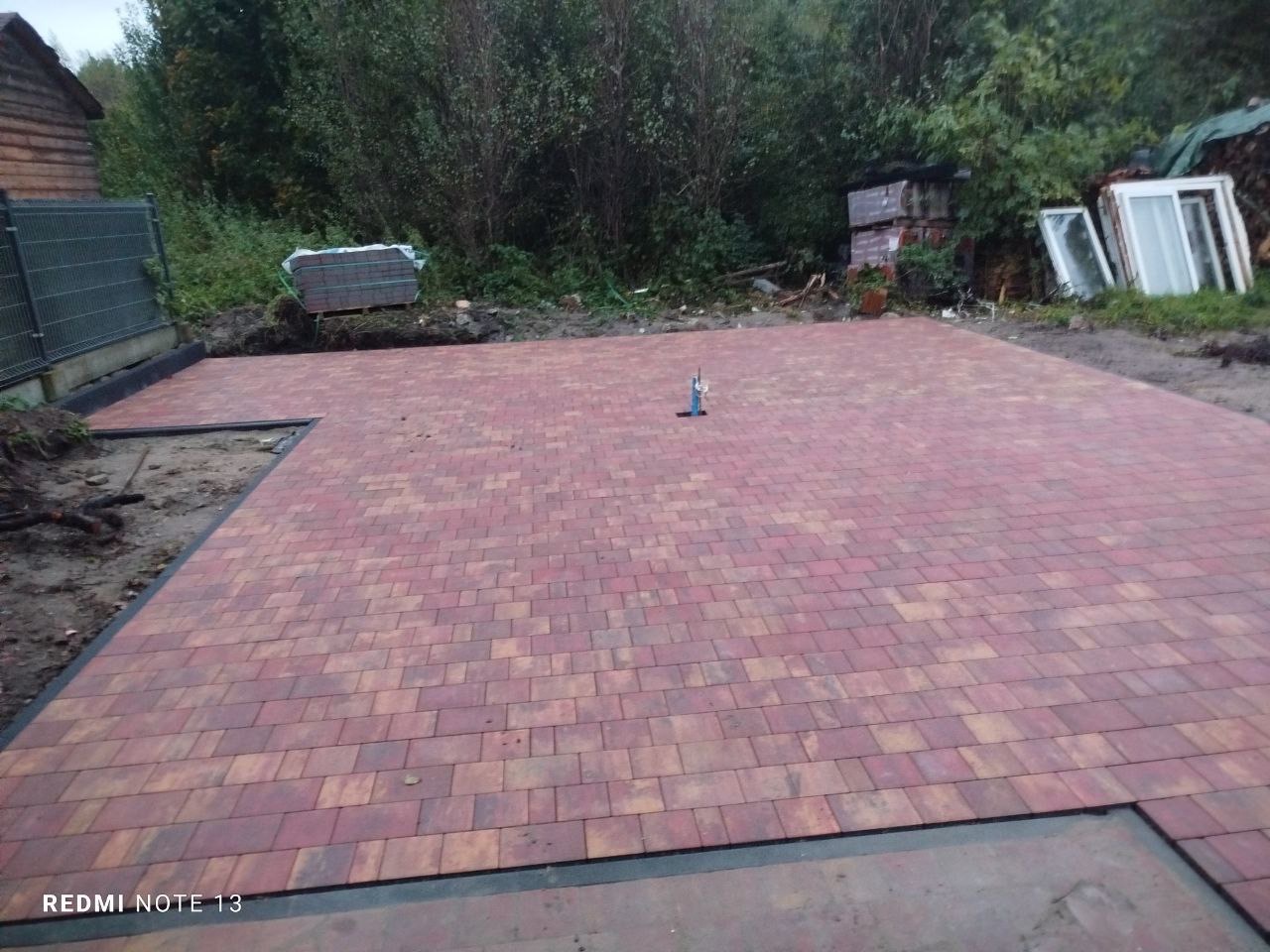 Ułożona, czerwono-brązowa kostka brukowa na posesji, z widocznym fragmentem ogrodzenia i stosem kostki w tle. Widoczne narzędzie do poziomowania.