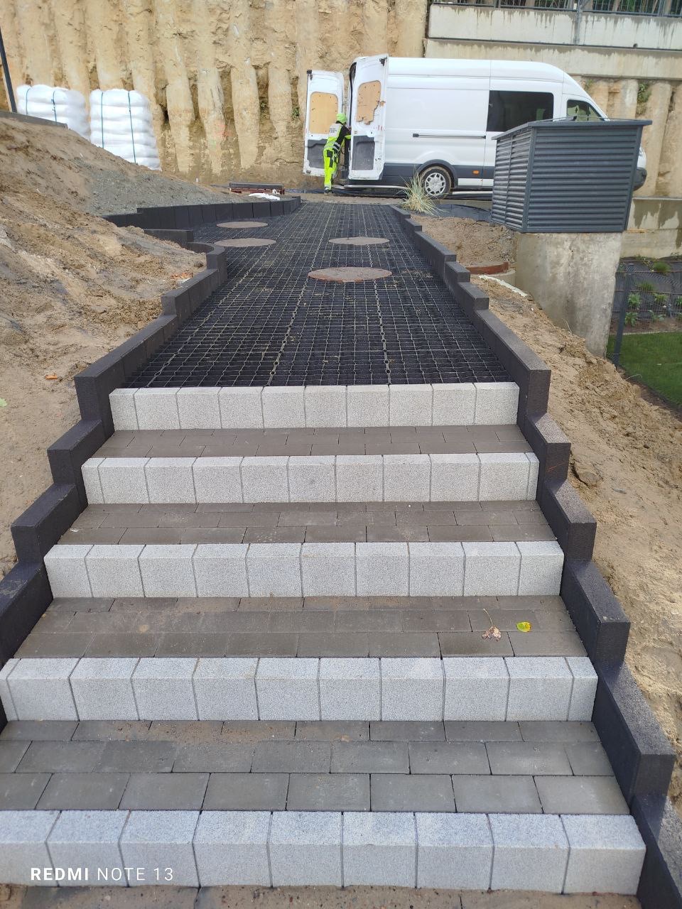 Schody z kostki brukowej prowadzące do kratki trawnikowej, z widocznym fragmentem terenu i pracującym przy samochodzie pracownikiem w tle.