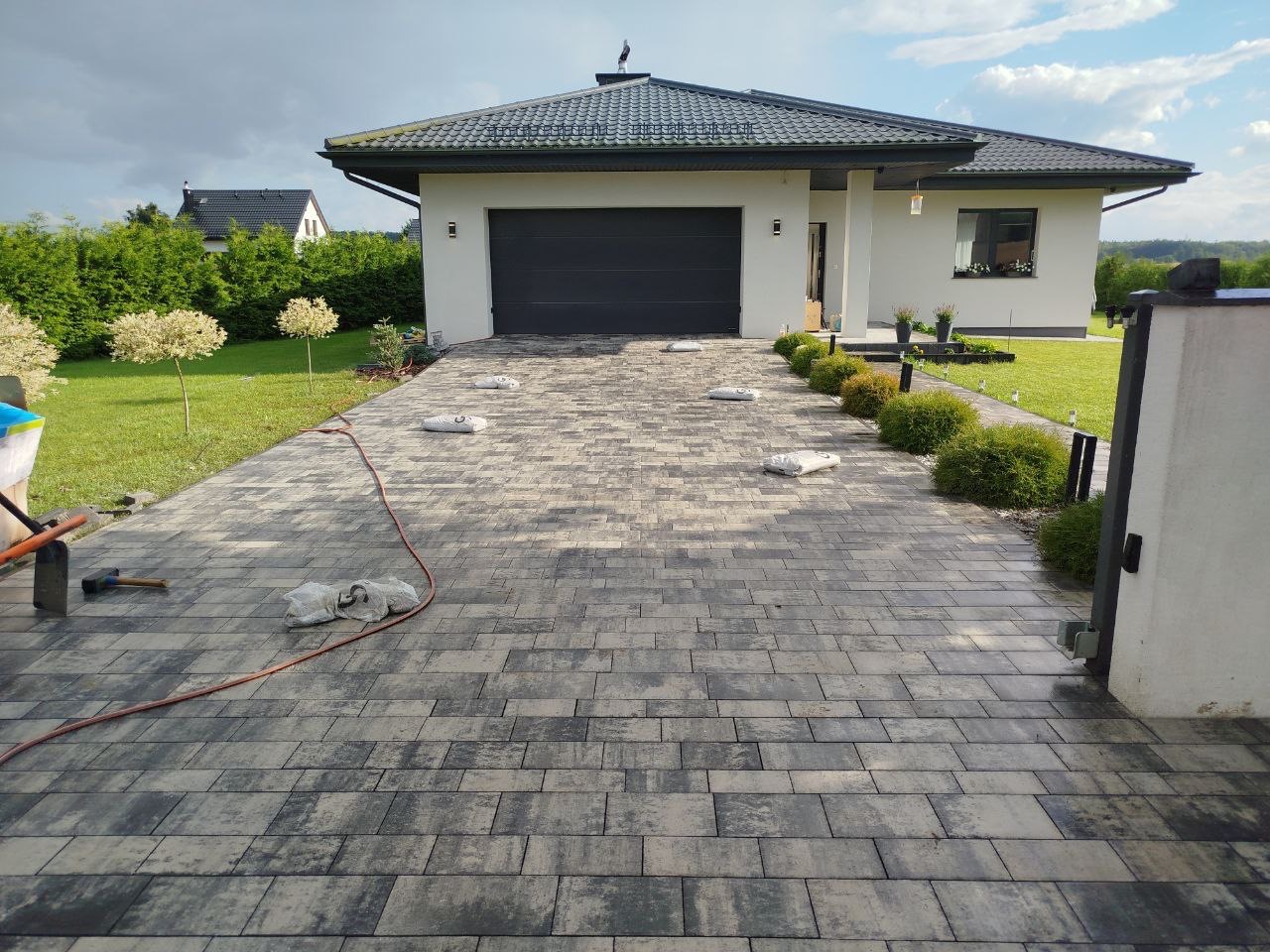 Ułożony podjazd z kostki brukowej w odcieniach szarości prowadzący do nowoczesnego domu z garażem. Widoczne worki z piaskiem i narzędzia brukarskie.