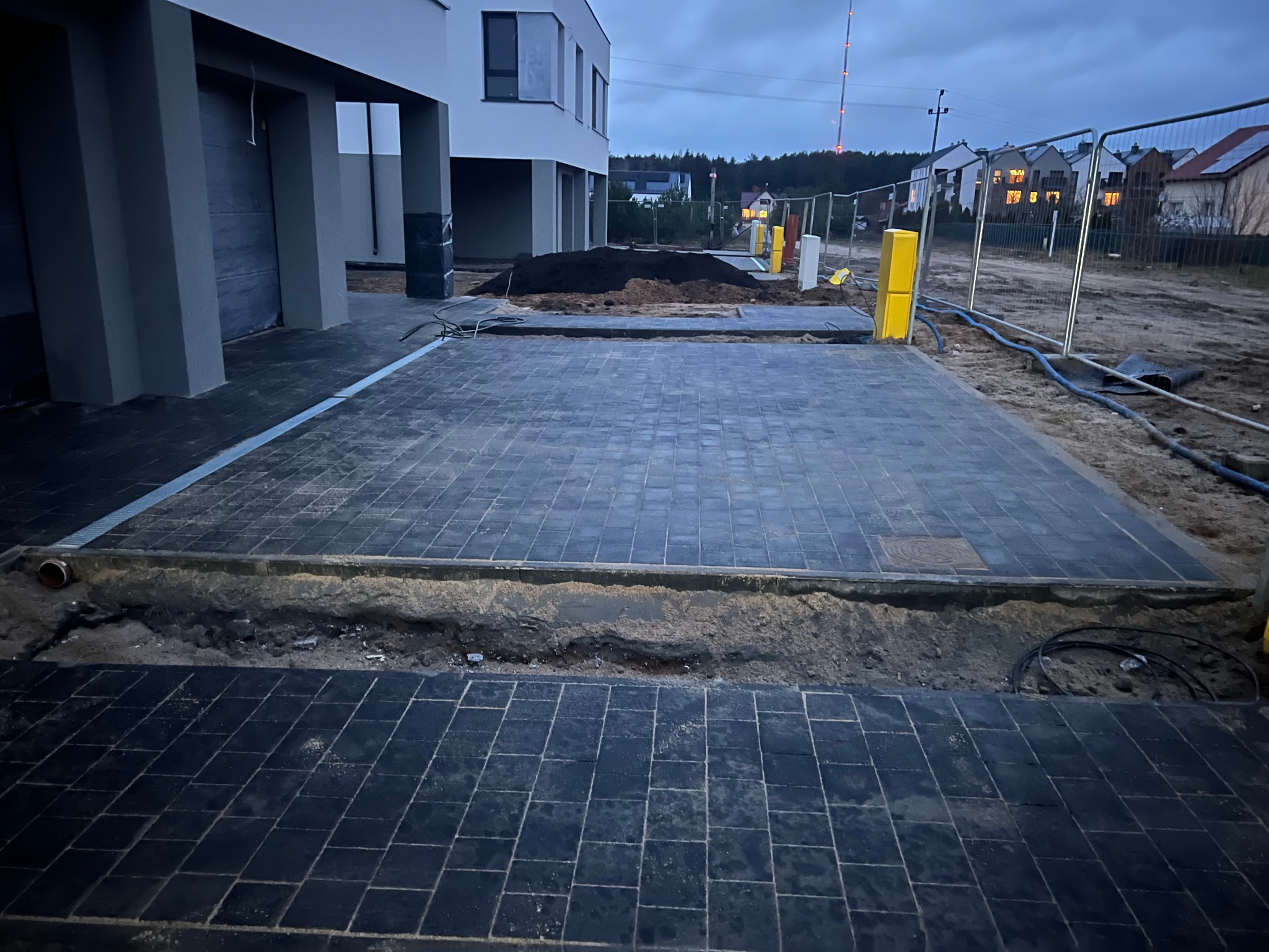 Ciemna kostka brukowa ułożona na podjeździe nowego domu w trakcie budowy. Widoczne wykopane obrzeża i przygotowany teren pod dalsze prace.