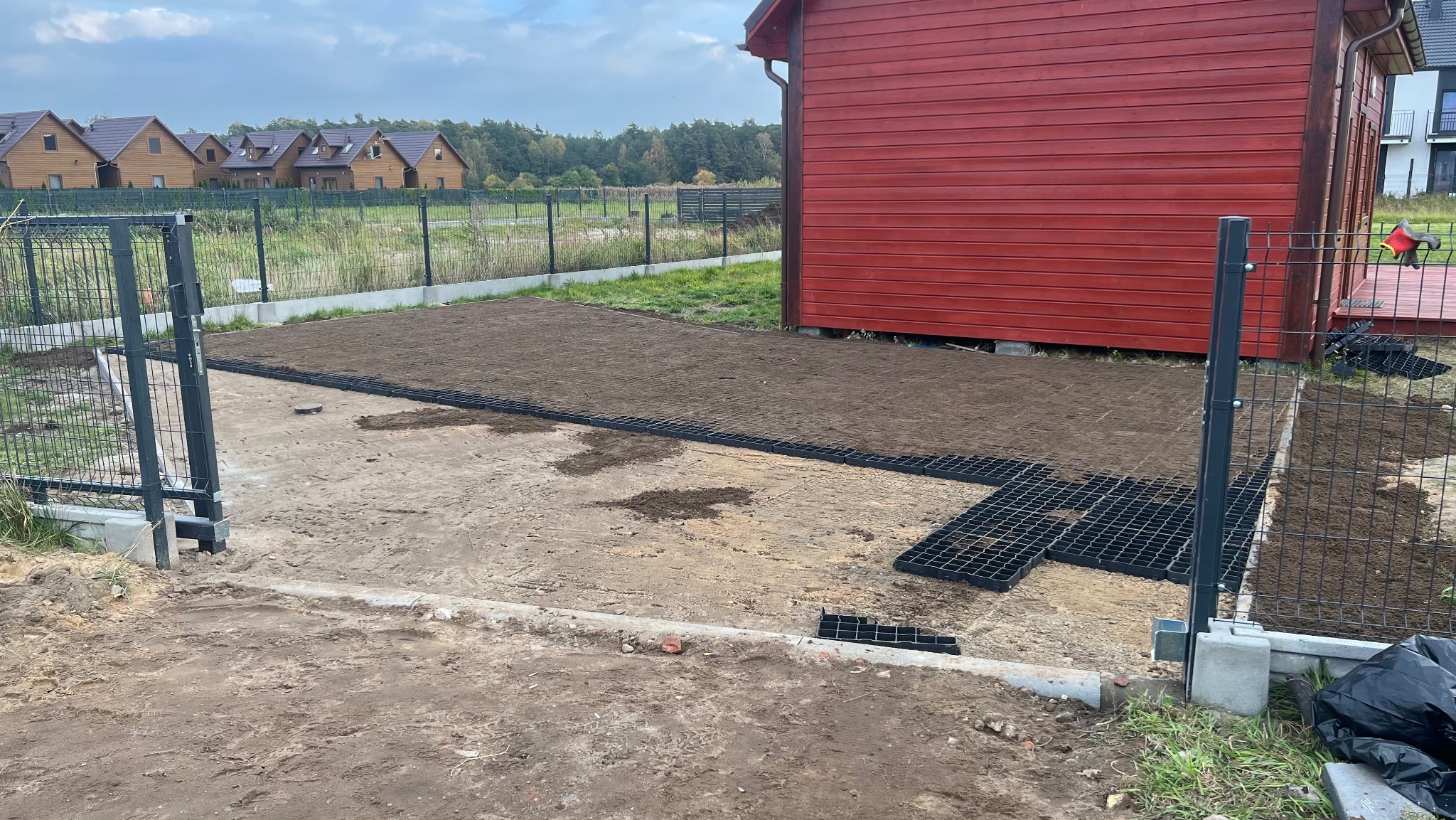 Przygotowanie terenu pod trawnik: wyrównana ziemia, siatka trawnikowa, ogrodzenie panelowe, czerwony budynek w tle, domy jednorodzinne w oddali.